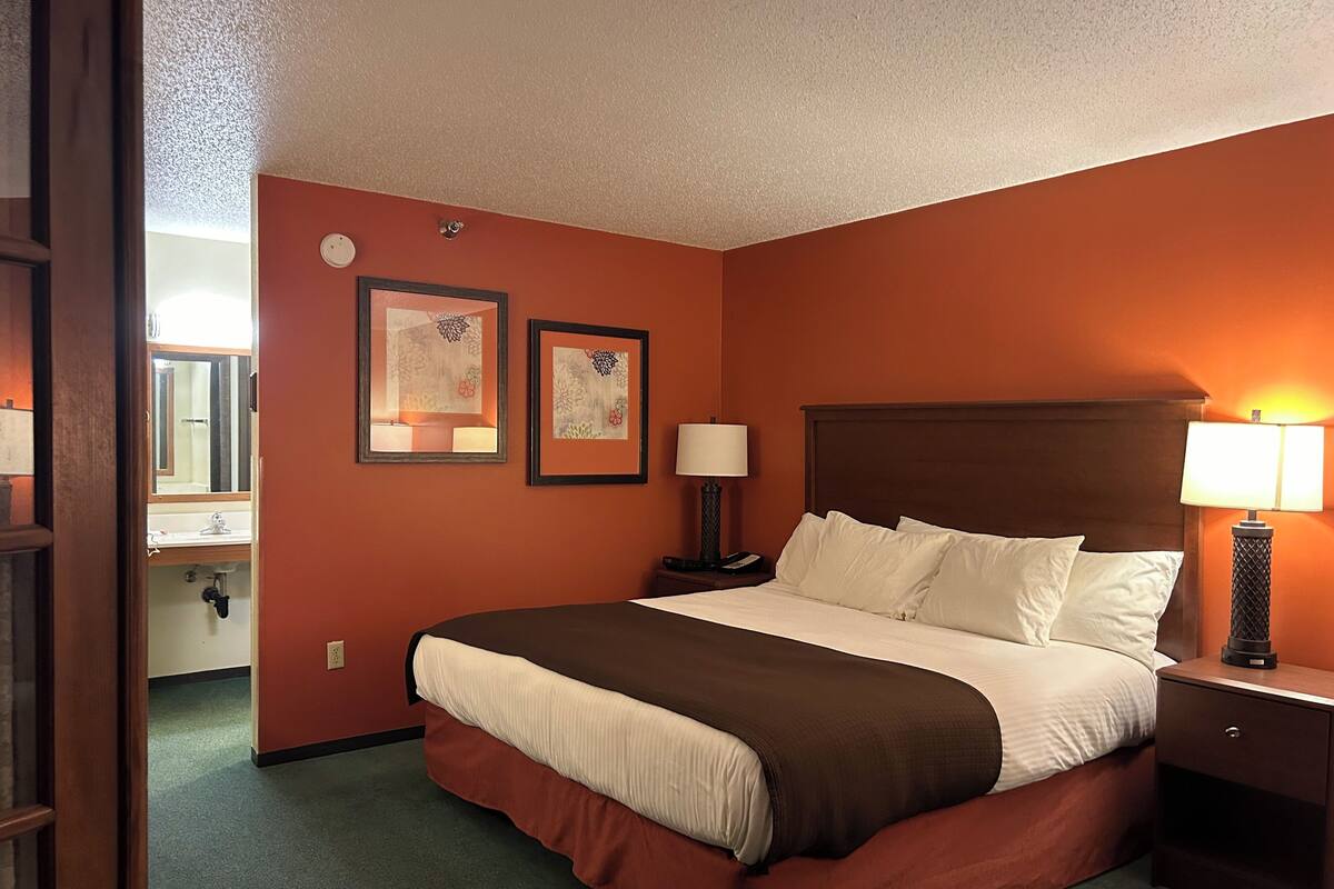 Quarto de hotel com uma cama grande, duas luminárias de cabeceira, um criado-mudo e um banheiro visível por uma porta aberta.