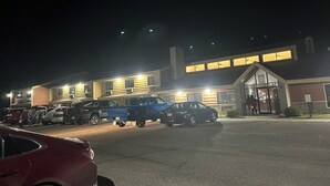 Een gebouw met een brede ingang, veel ramen en een parkeerplaats met verschillende voertuigen.