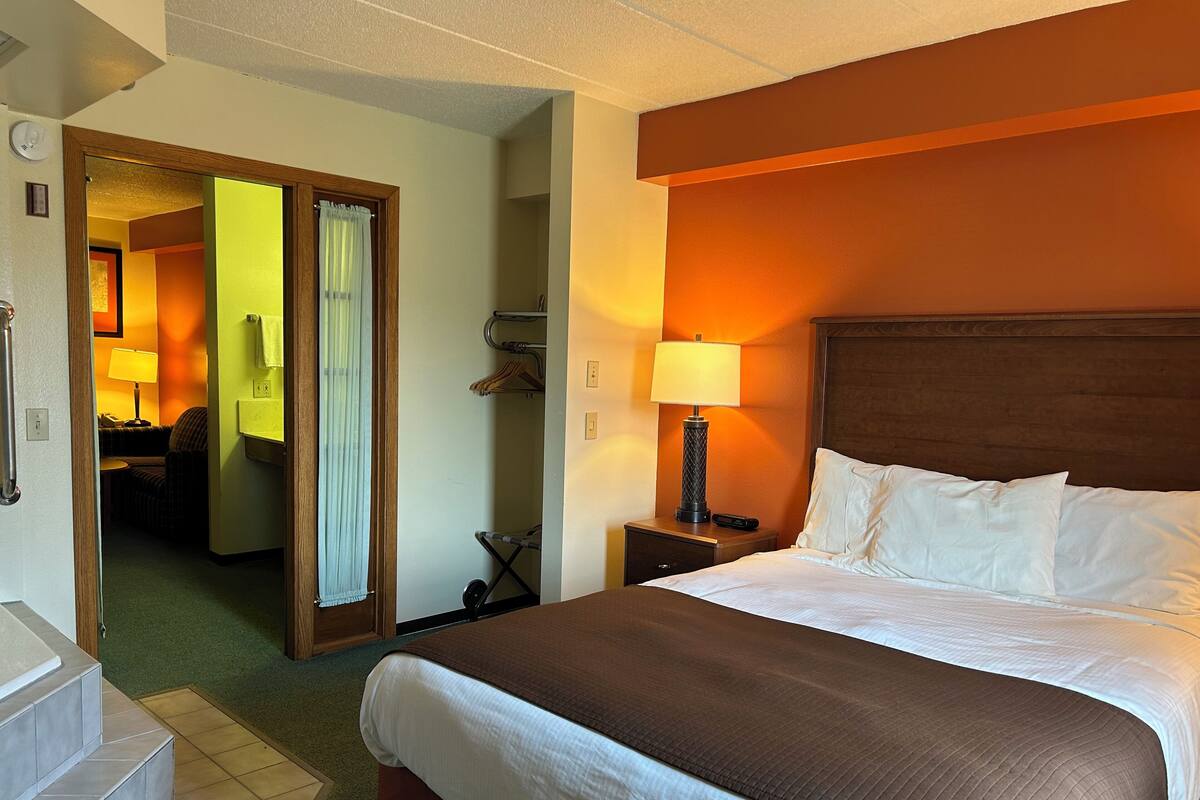Quarto de hotel com cama grande, mesa de cabeceira com abajur, banheiro com cortina de chuveiro e corredor que dá para outro quarto.