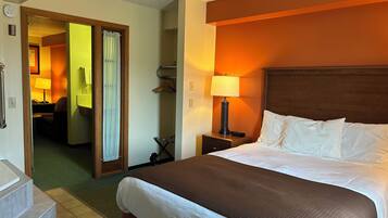 Une chambre d’hôtel avec un grand lit, une table de chevet avec une lampe, une salle de bain avec un rideau de douche et un couloir menant à une autre chambre.