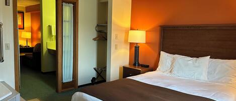 Una habitación de hotel con una cama grande, una mesita de noche con lámpara, un baño con cortina de ducha y un pasillo que conduce a otra habitación.