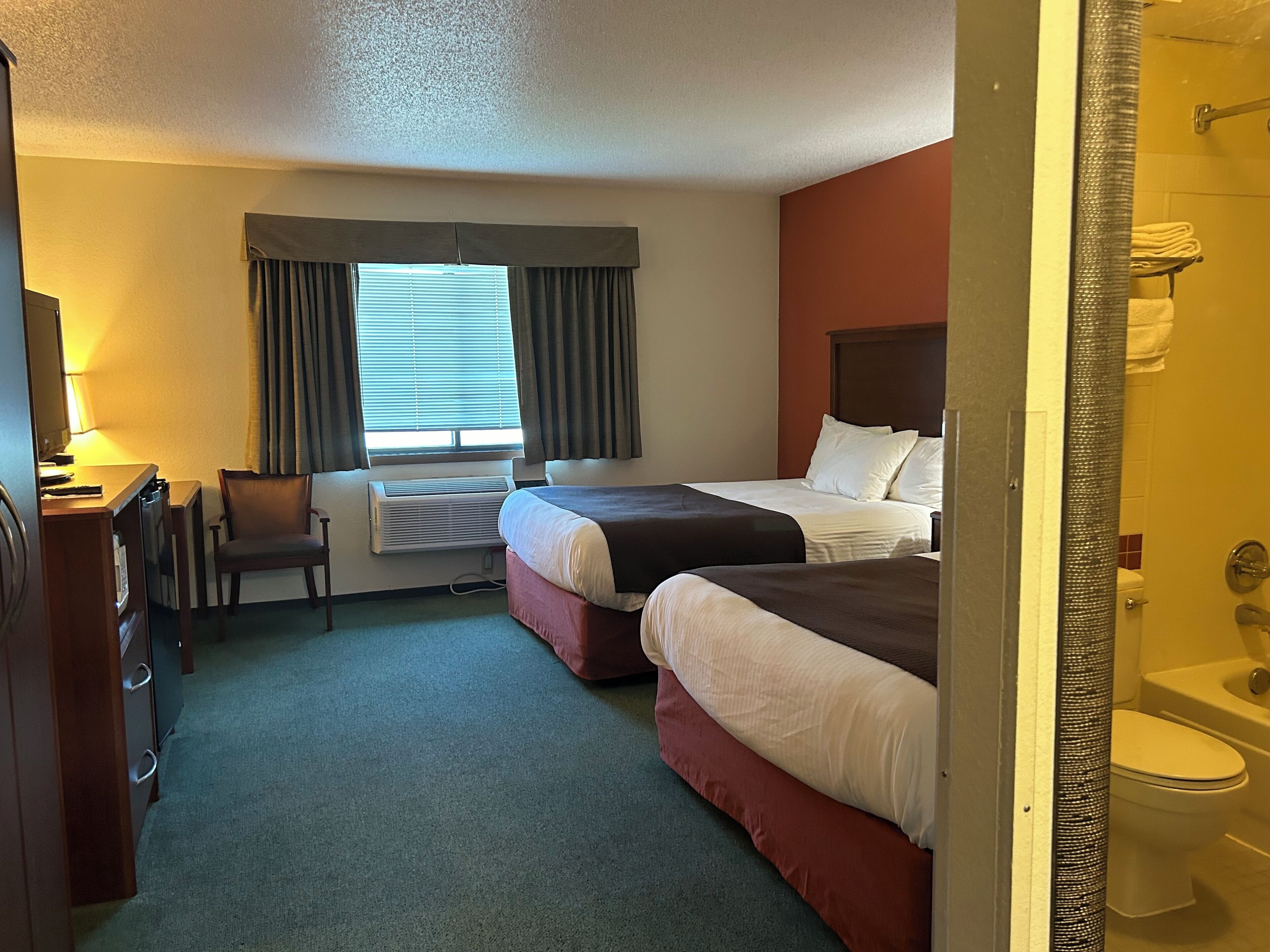 A hotel room with two beds, a desk with a chair, a window with curtains, and a bathroom visible through an open door.