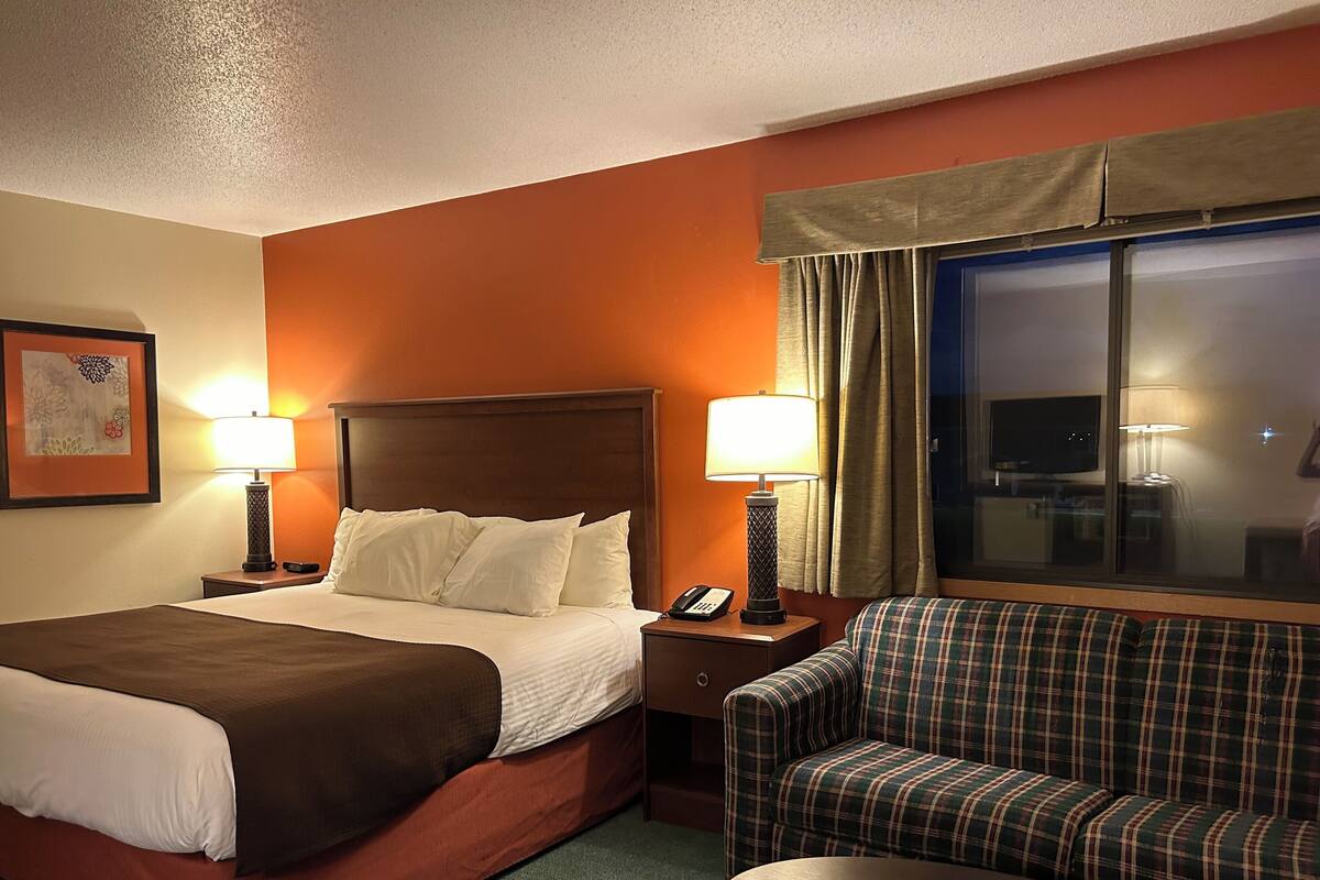 Quarto de hotel com uma cama, duas luminárias de cabeceira, uma cadeira, uma mesinha e uma janela com cortinas.