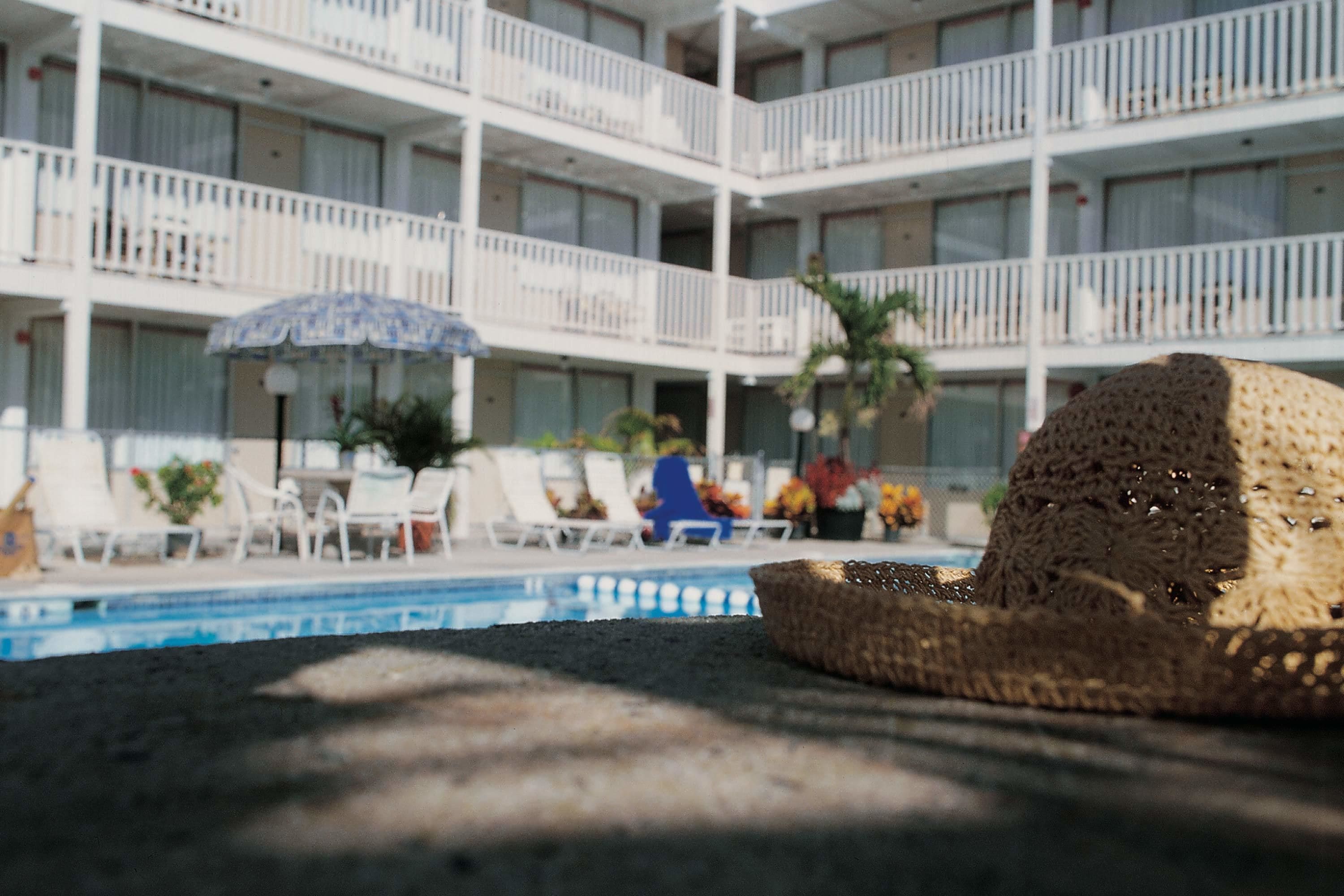 Una sombrilla de playa, sillas de descanso y una zona junto a la piscina, con un edificio al fondo.