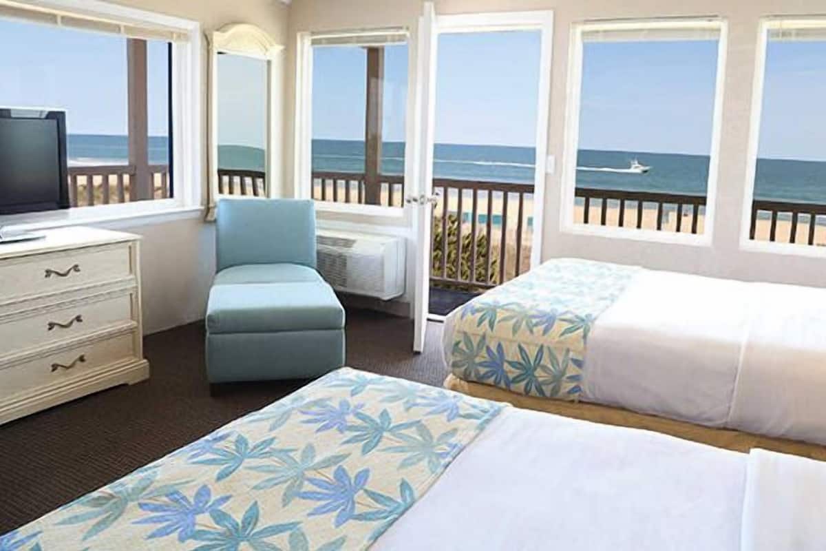 A hotel room with two beds, a TV, a chair, and a view of the ocean.