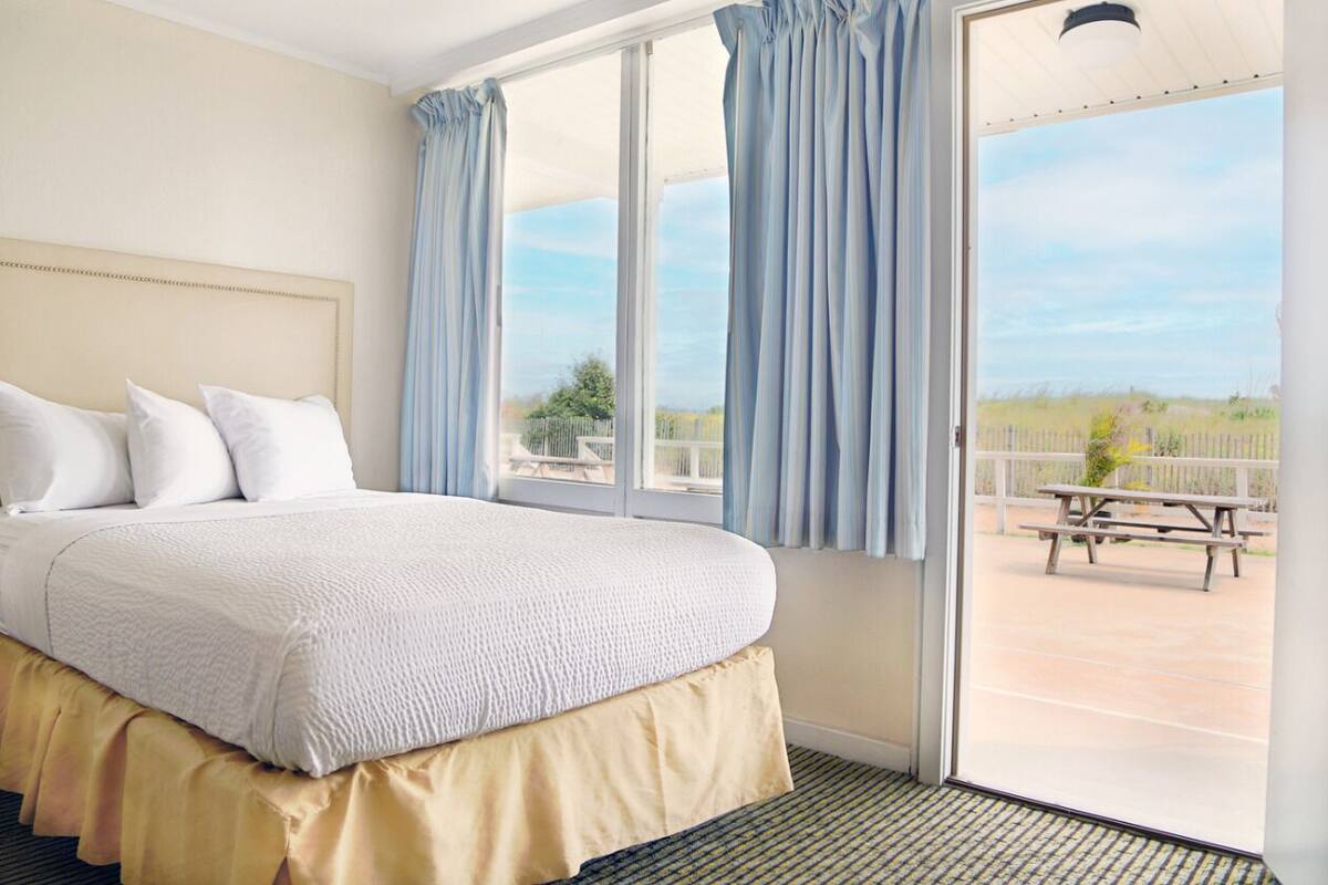 A bedroom with a large bed, a view of a patio with a picnic table, and a balcony with blue curtains.
