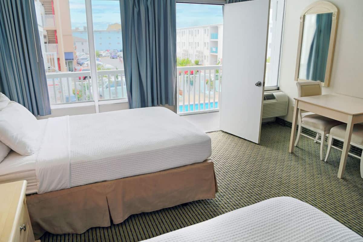 A hotel room with a bed, a desk, a chair, and a large window with a view of buildings and a pool.