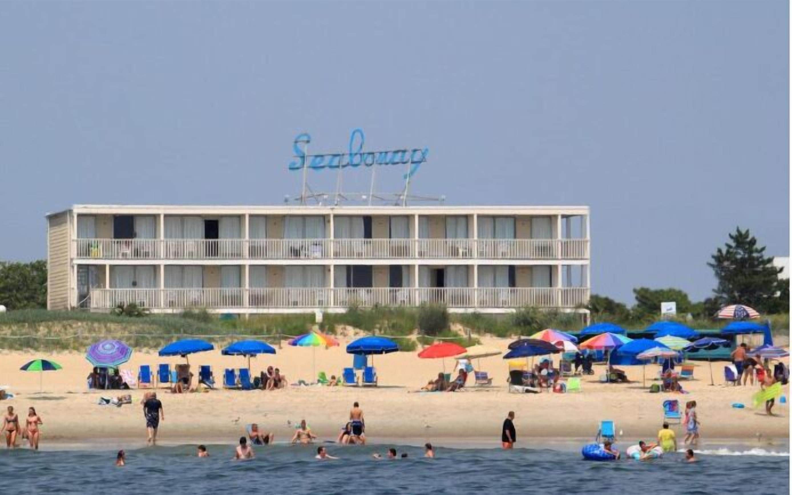 A hotel with a balcony, located on a beach with umbrellas and people.