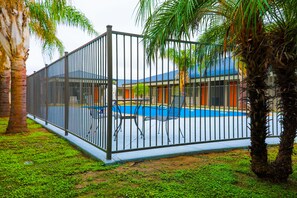 A fenced pool area with a blue pool, palm trees, and a building with a blue roof in the background.