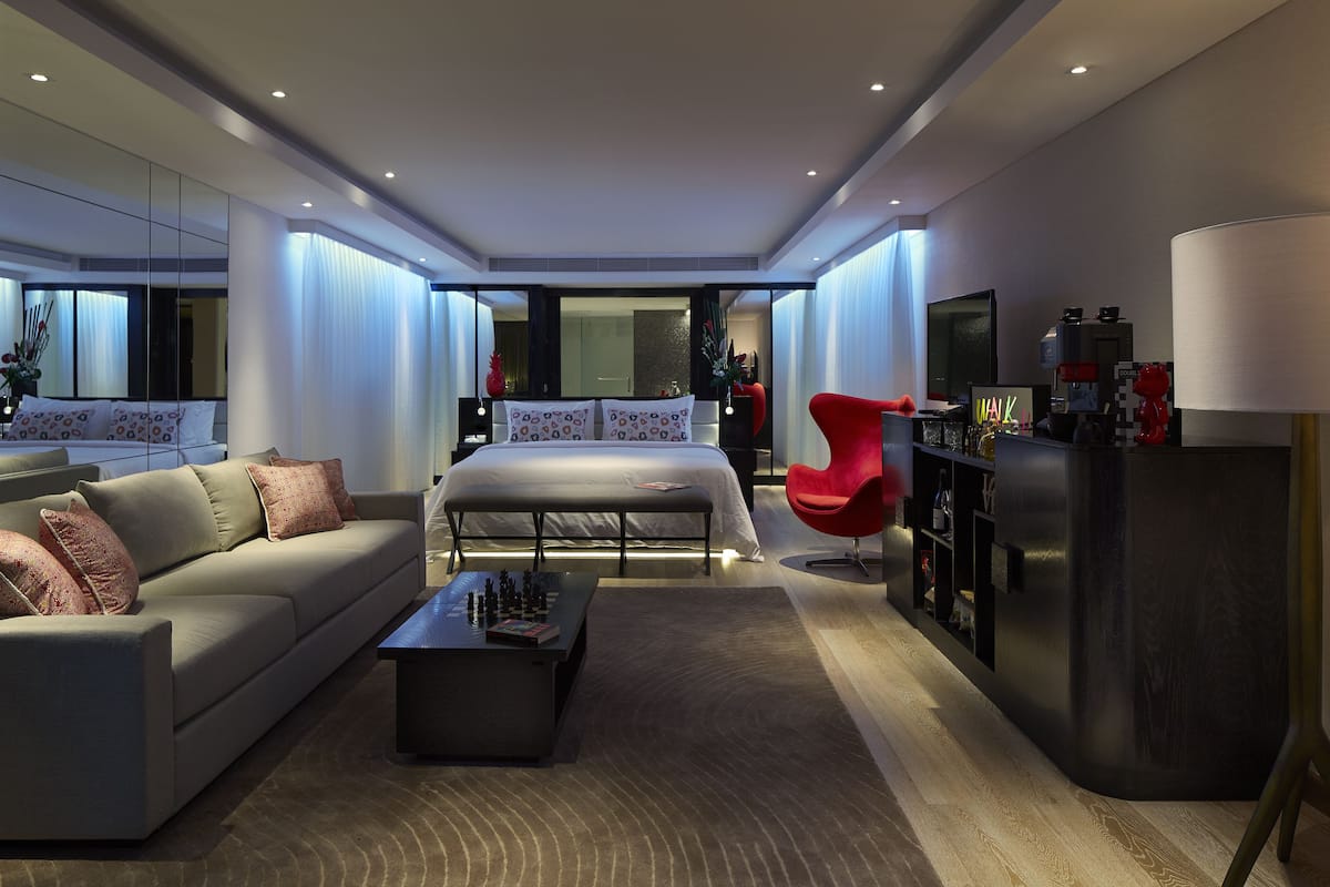 A modern hotel room with a sofa, a bed, a red chair, a coffee table, and a TV.