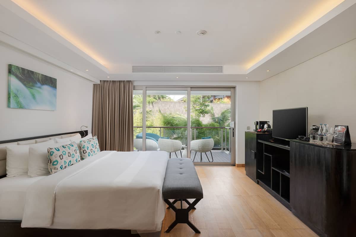 A modern hotel room with a large bed, a bench, a TV, and a balcony with outdoor seating.