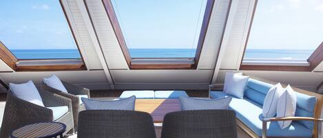 A sunlit outdoor seating area with a view of the sea, featuring wicker furniture and a wooden table.