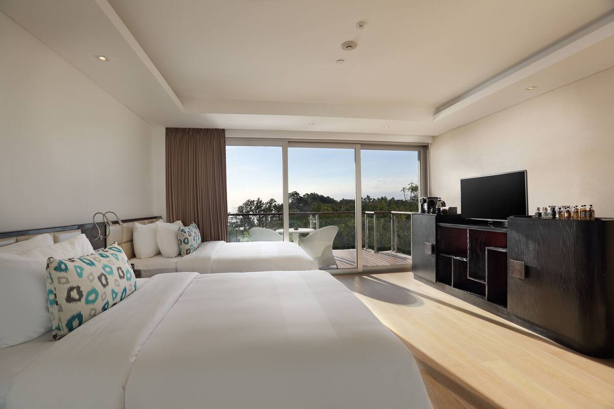 A modern hotel room with two beds, a TV, a balcony with a table and chairs, and a wooden cabinet.