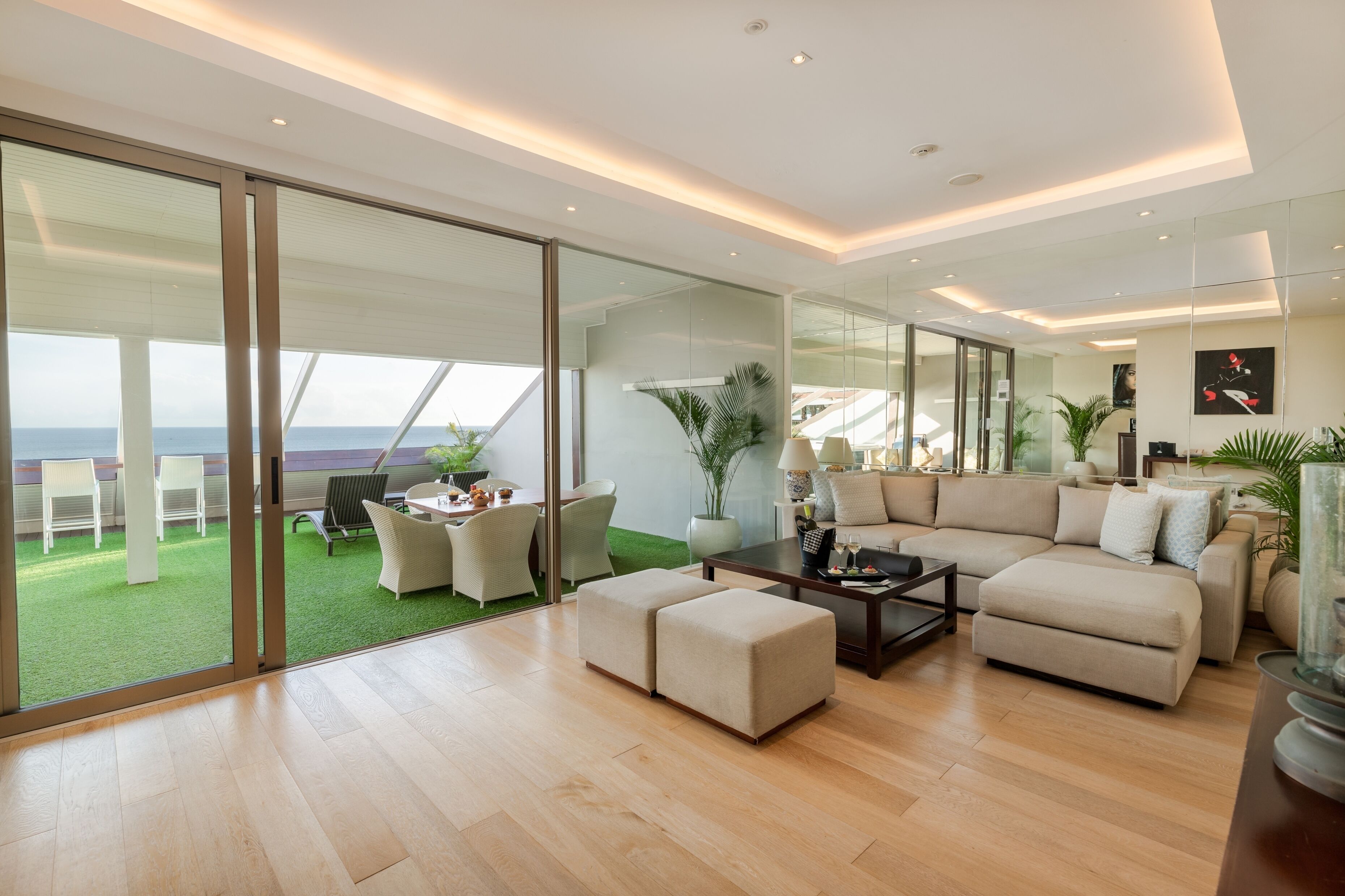 A modern living room with a large glass door leading to a balcony with a view of the sea. There is a beige sofa, a coffee table, and a dining area with chairs.