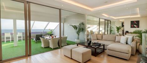 A modern living room with a large glass door leading to a balcony with a view of the sea. There is a beige sofa, a coffee table, and a dining area with chairs.