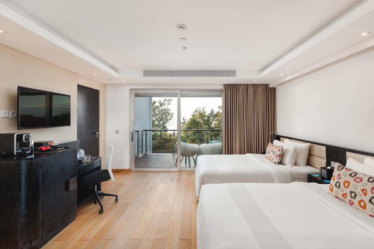 A hotel room with two beds, a desk with a flat-screen TV, a chair, and a balcony with a view of trees.