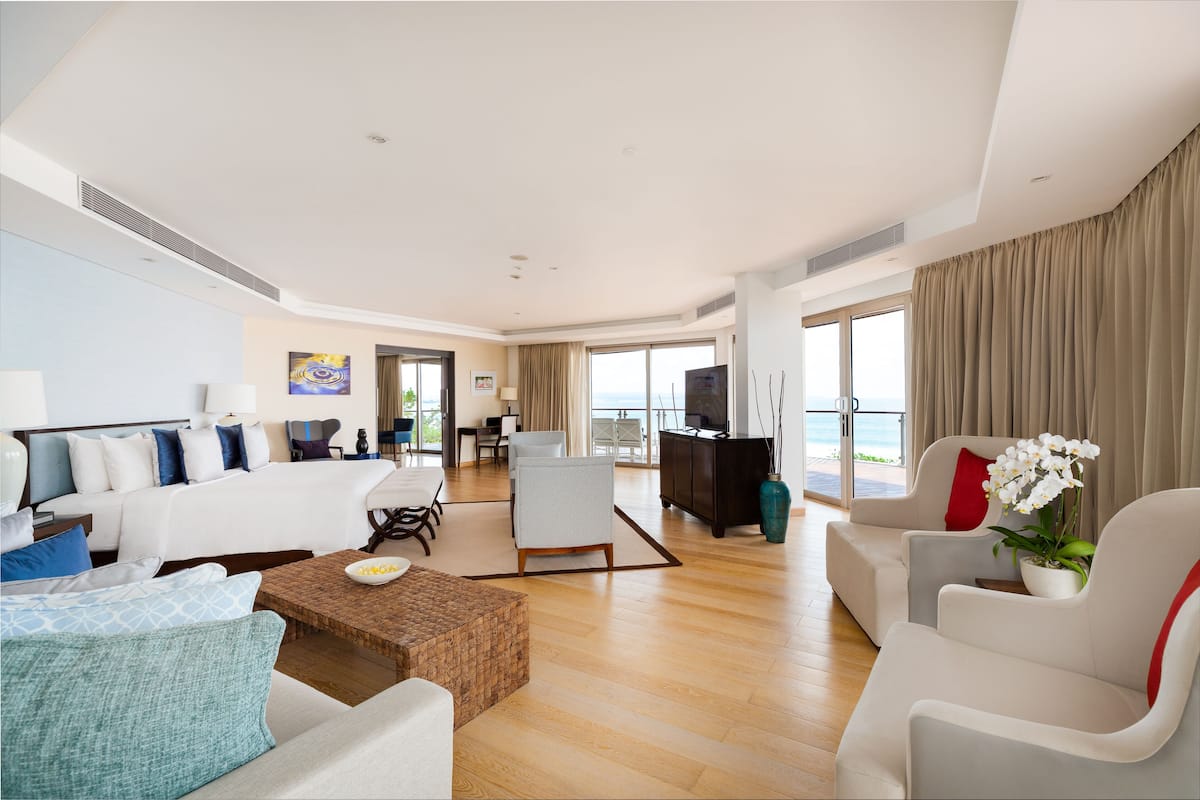A spacious hotel room with a large bed, a sitting area with a sofa and armchairs, a dining table, and a view of the ocean.