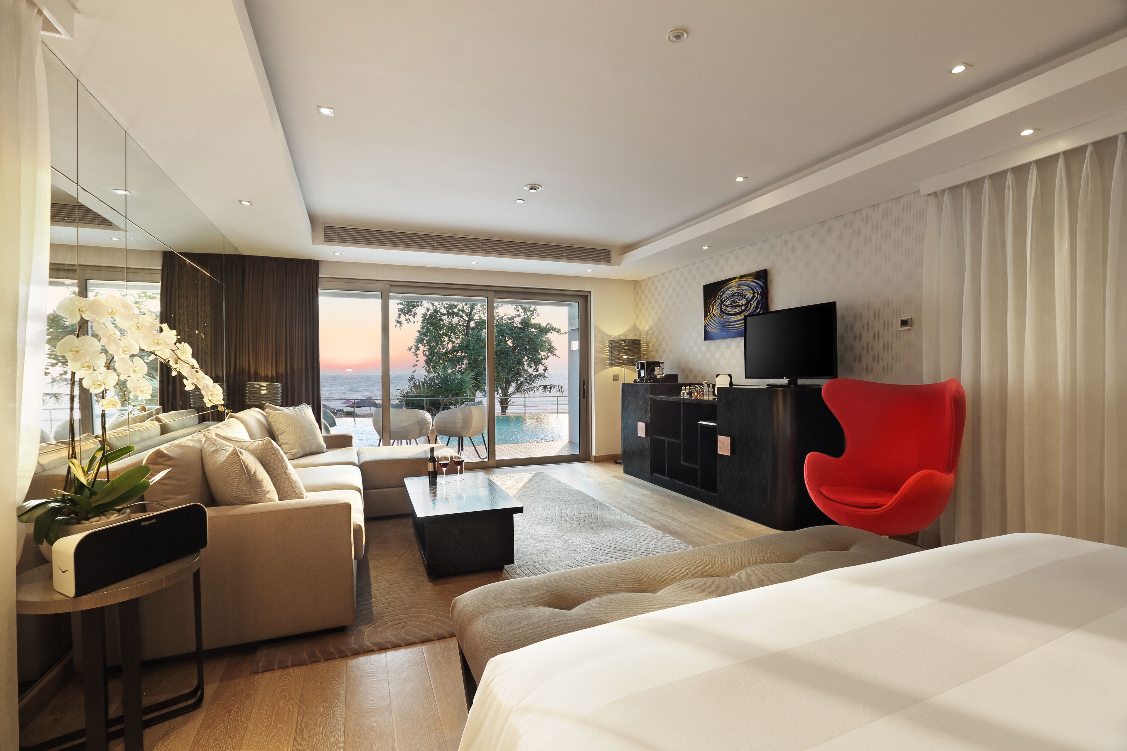 A modern hotel room with a large bed, a sofa, a red chair, a TV, and a view of the outdoors.