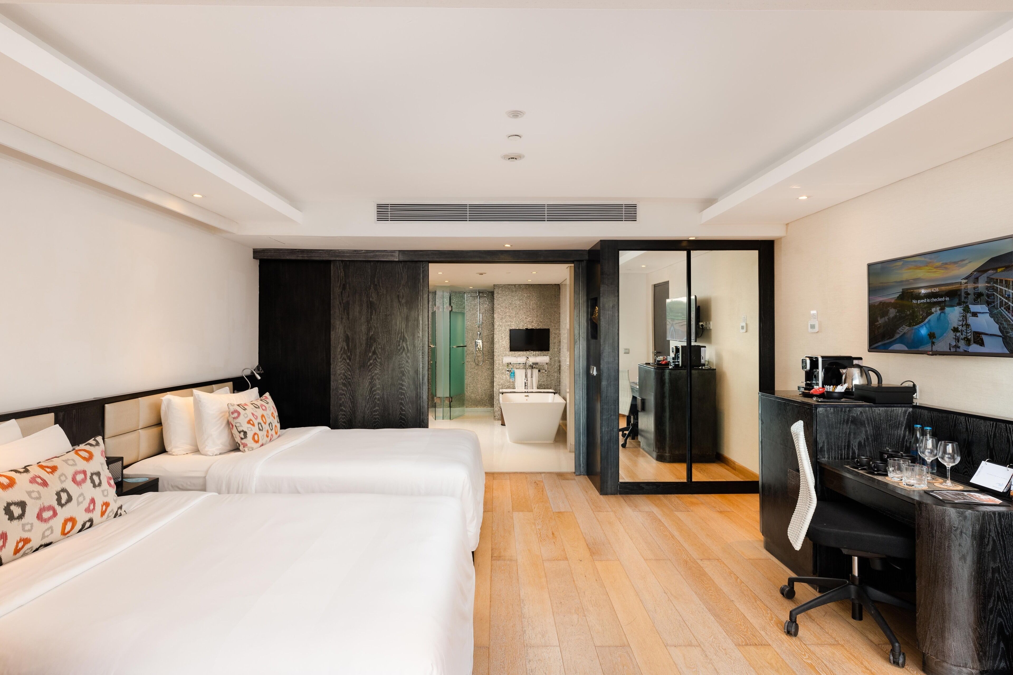 Quarto de hotel moderno com duas camas, uma escrivaninha com cadeira, um quadro grande e um banheiro visível através de uma porta aberta.