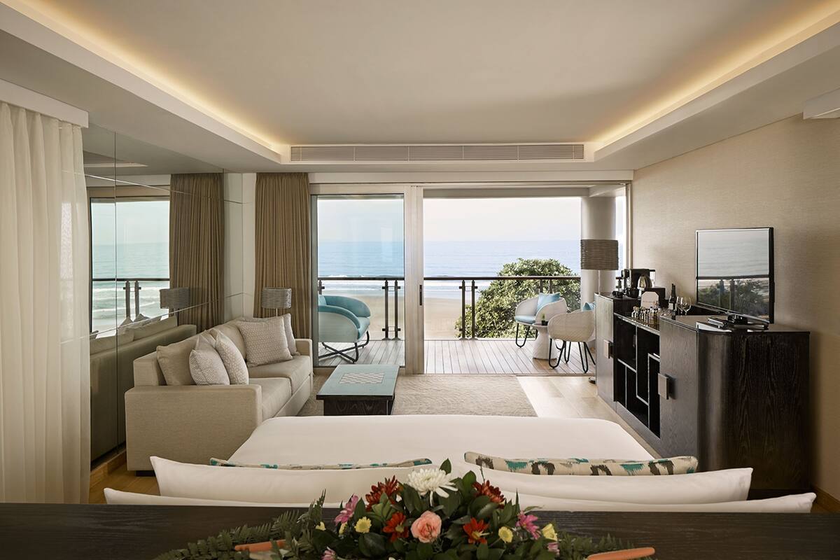 A hotel room with a large bed, a sofa, a desk, and a TV. There is a balcony with a view of the ocean.