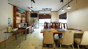 A dining area with a long table, chairs, a television, and a shelf with various items.