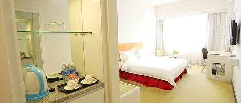 A hotel room with a bed, a desk, a chair, a TV, a mirror, and a kitchenette area with a kettle and tea set.