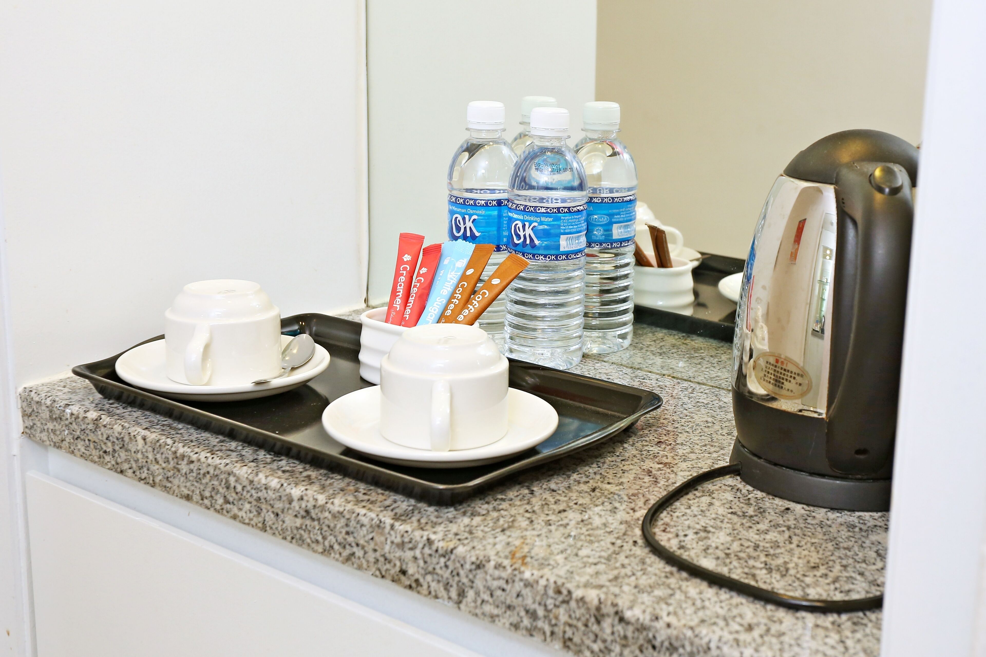 A kitchen counter with a coffee maker, two white cups, a tray with utensils, and several water bottles.