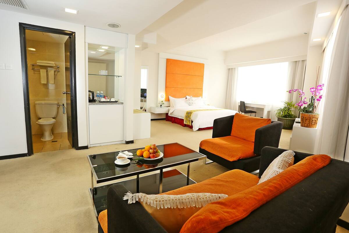 A hotel room with a bed, a sitting area with a sofa and armchair, a small table with fruit, a bathroom with a toilet and sink, and a large window.