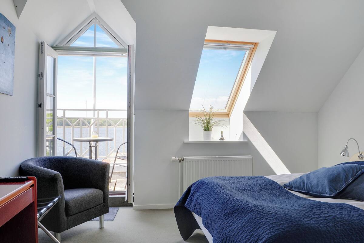 A bedroom with a skylight, a bed with blue bedding, a chair, and a view of the outdoors.