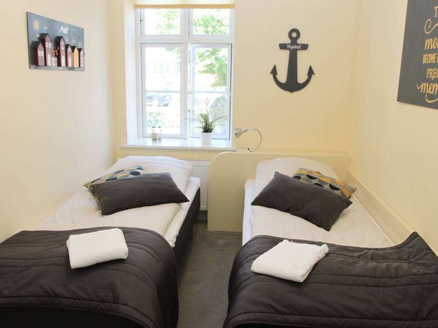 A room with two beds, a window with a view, a plant on the windowsill, and a wall with a nautical-themed decoration.