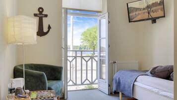 Una habitación con una cama, una silla verde, una mesa con una lámpara, un cuadro y vistas a través de una puerta abierta.