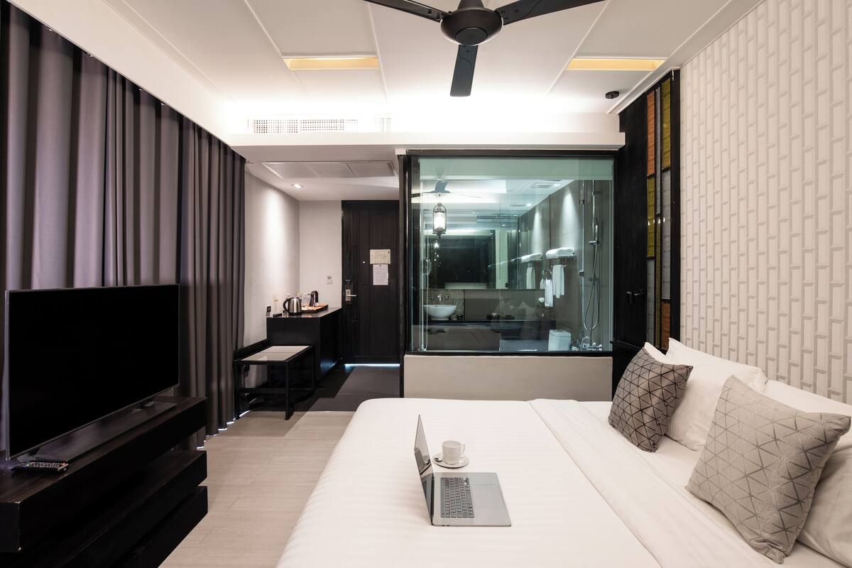 A modern hotel room with a flat-screen TV, a laptop on the bed, a desk with a chair, and a large window with a view of the bathroom.