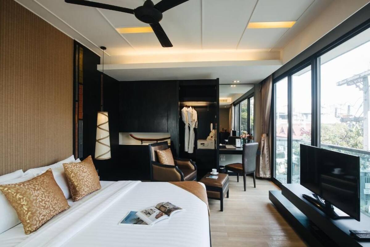 A modern hotel room with a large bed, a desk, a chair, a television, and a balcony with a view.
