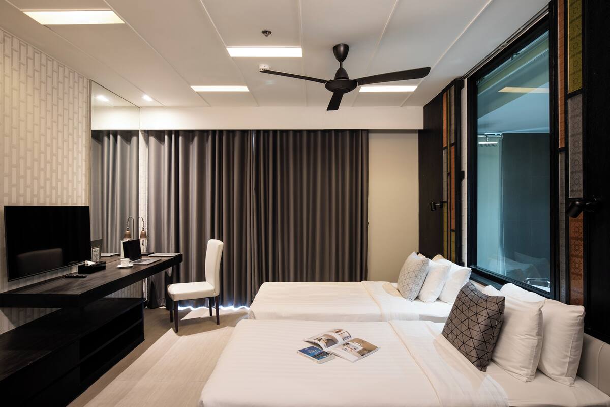 A hotel room with a flat-screen TV, a desk with a chair, two beds with pillows, a ceiling fan, and a large window.