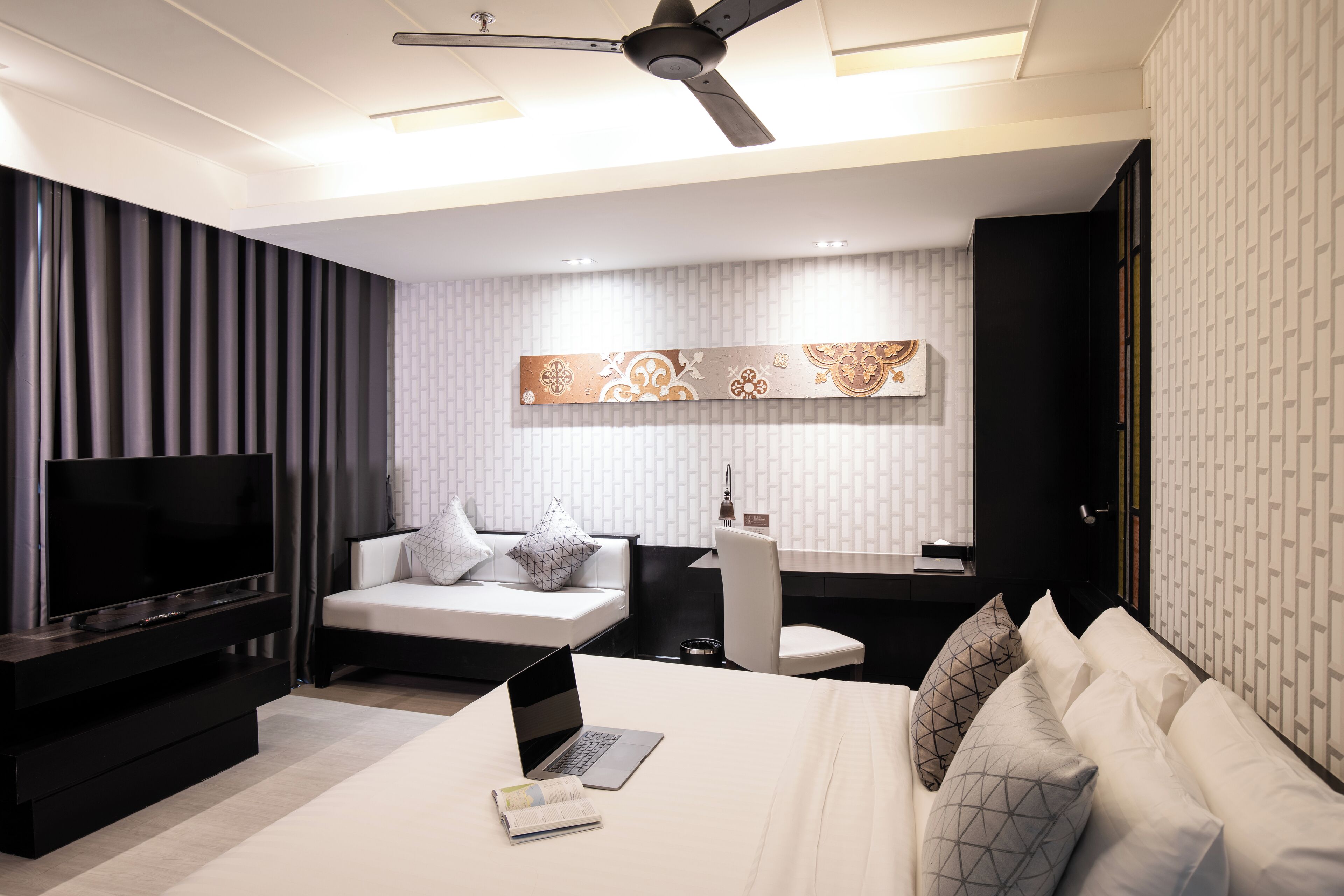 Quarto moderno com televisão de tela plana, um laptop, cama com roupa de cama branca, escrivaninha com cadeira e ventilador de teto.