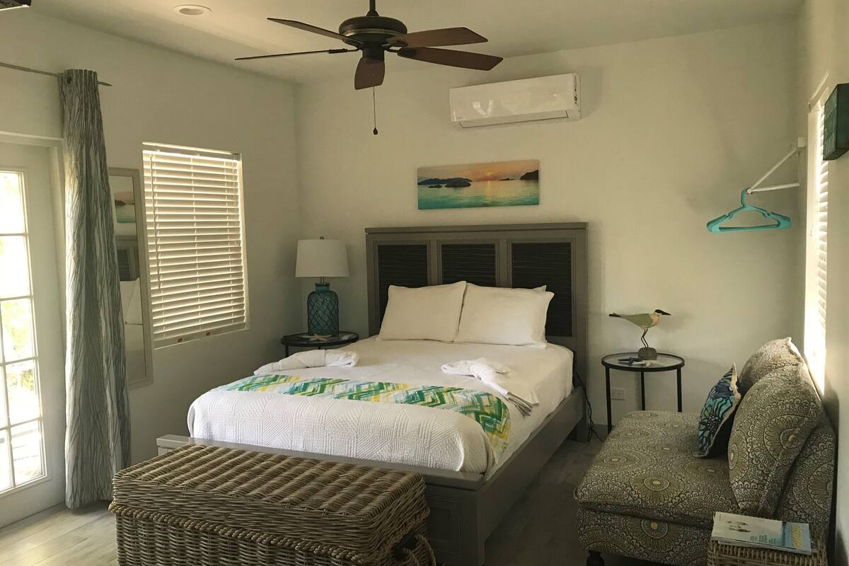 A bedroom with a bed, a ceiling fan, a nightstand, a lamp, a chair, a hanging clothes rack, and a painting on the wall.