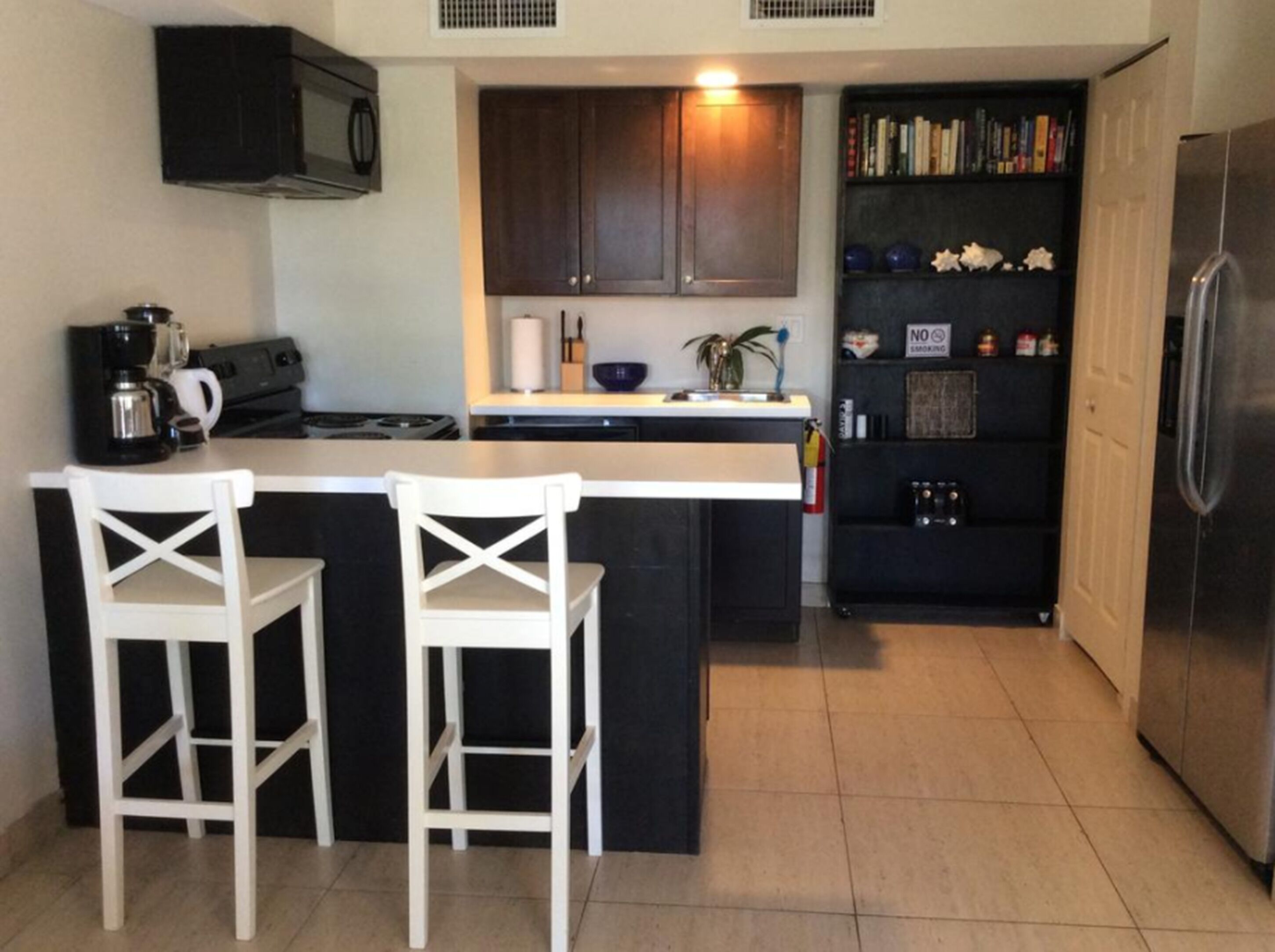 Una cocina compacta con una isla central, dos taburetes blancos, armarios oscuros, un microondas, un refrigerador y una estantería con libros y objetos decorativos.