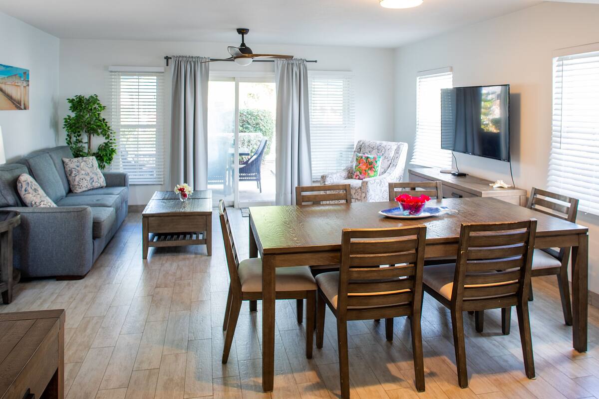 A modern living room with a dining area, including a wooden table and chairs, a sofa, and a television.