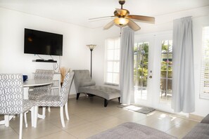 A modern living room with a flat-screen TV, a ceiling fan, a dining area with patterned chairs, and large windows with curtains.