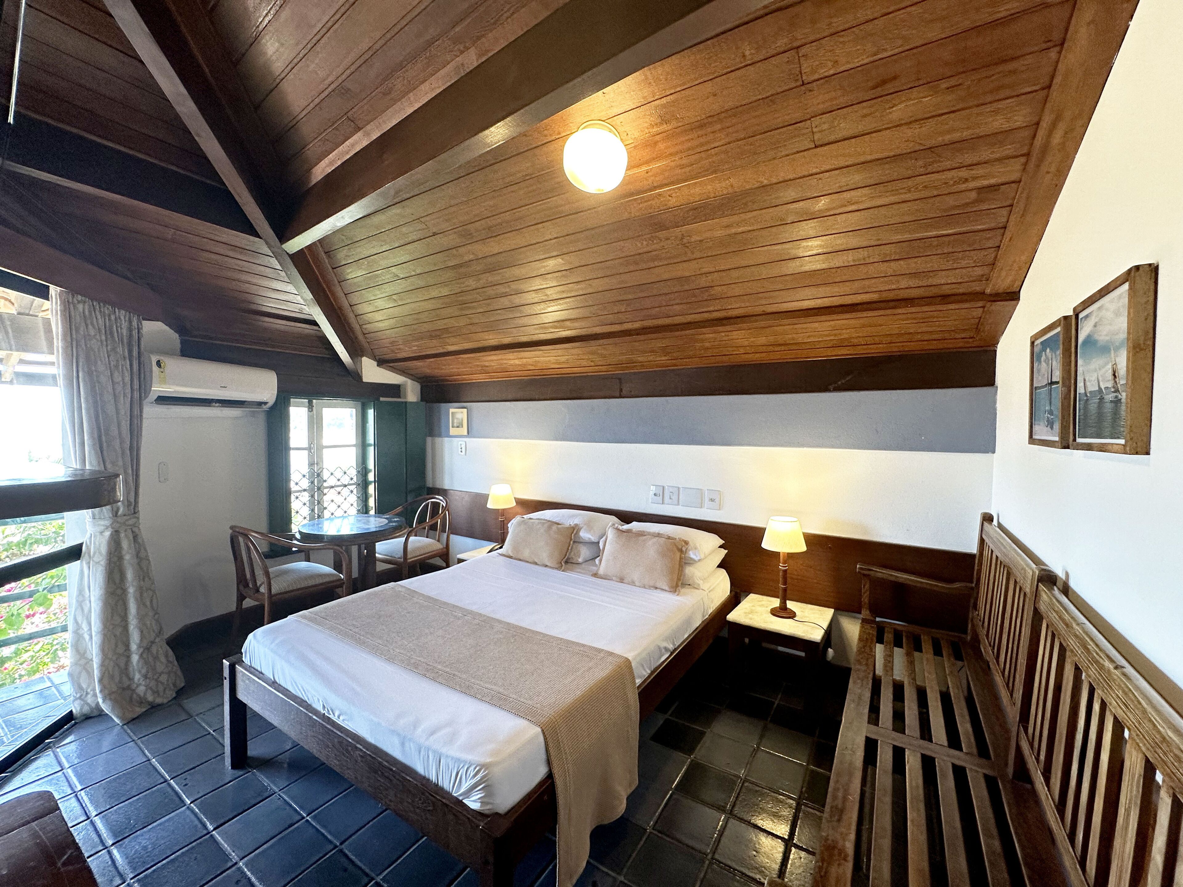 A room with a large bed, two bedside tables with lamps, a wooden ceiling, and a window with curtains.