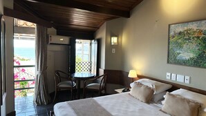 Habitación de hotel con cama, una mesa con sillas, un cuadro en la pared y vista al mar a través de una ventana.