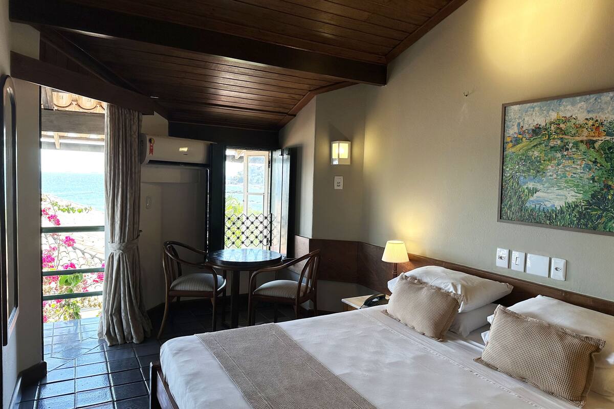 Habitación de hotel con cama, una mesa con sillas, un cuadro en la pared y vista al mar a través de una ventana.