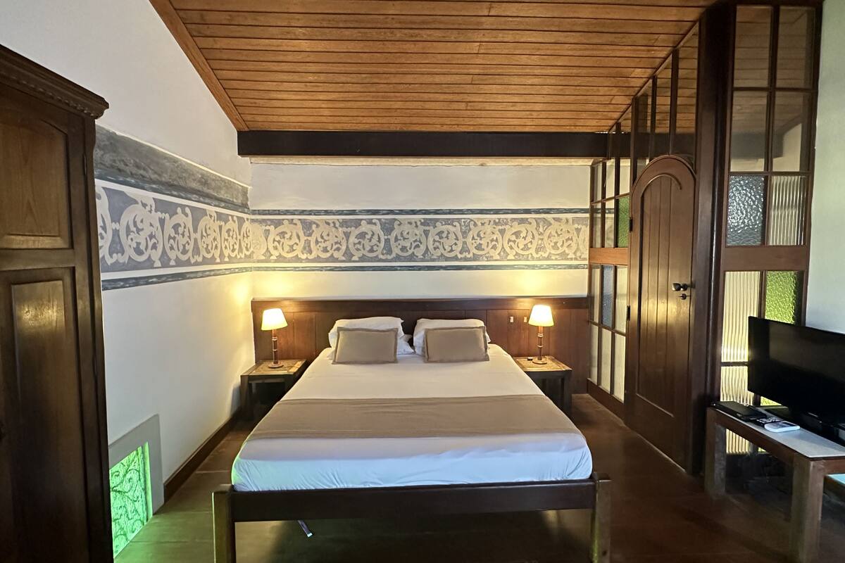 Un dormitorio con techo de madera, una cama con dos almohadas, mesitas de noche con lámparas, un televisor sobre un mueble bajo y un armario.