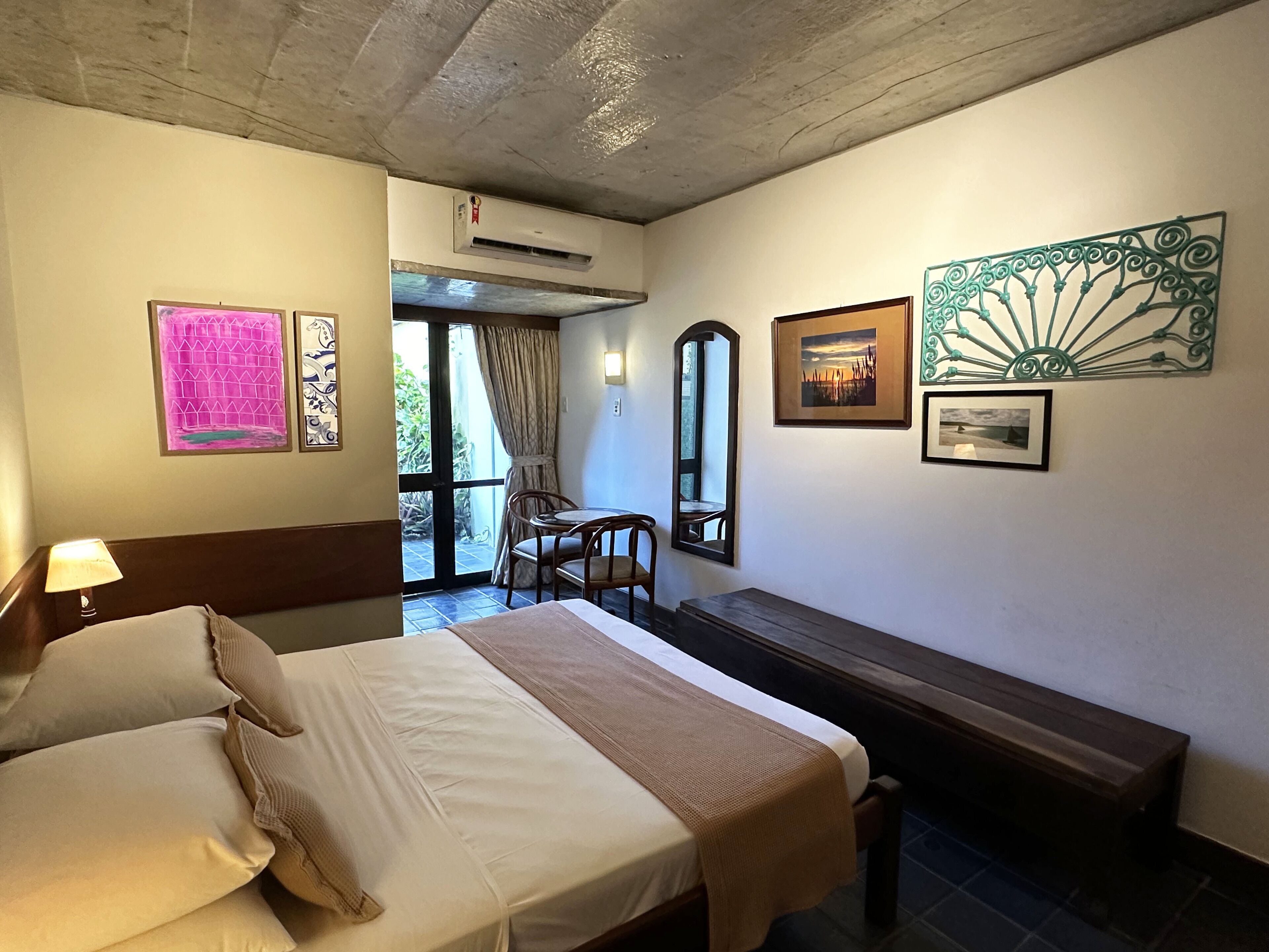 A hotel room with a bed, a bench, a table, and chairs. There are pictures on the wall, an air conditioning unit, and a view of the outside through a door.