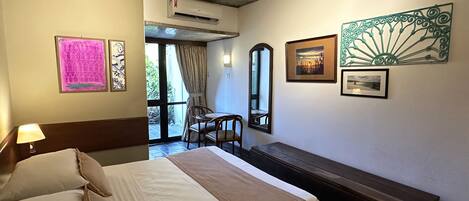 A hotel room with a bed, a bench, a table, and chairs. There are pictures on the wall, an air conditioning unit, and a view of the outside through a door.