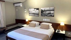 Habitación de hotel con una cama grande, dos mesitas de noche con lámparas, aire acondicionado de pared y dos fotografías en blanco y negro.