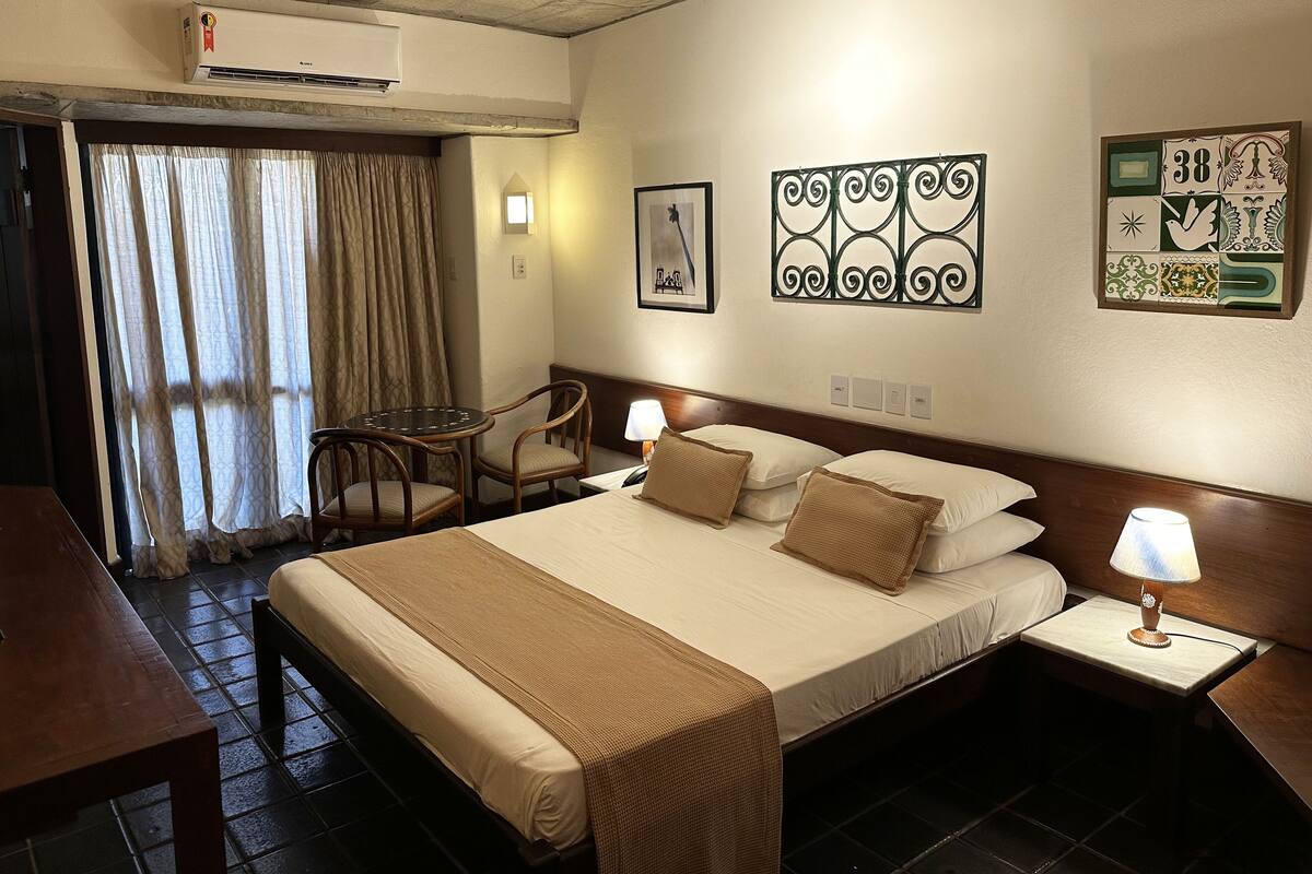 Habitación de hotel con cama, dos sillas, una mesita y un escritorio. Se observa una unidad de aire acondicionado en el techo, una ventana con cortinas y decoración en la pared.
