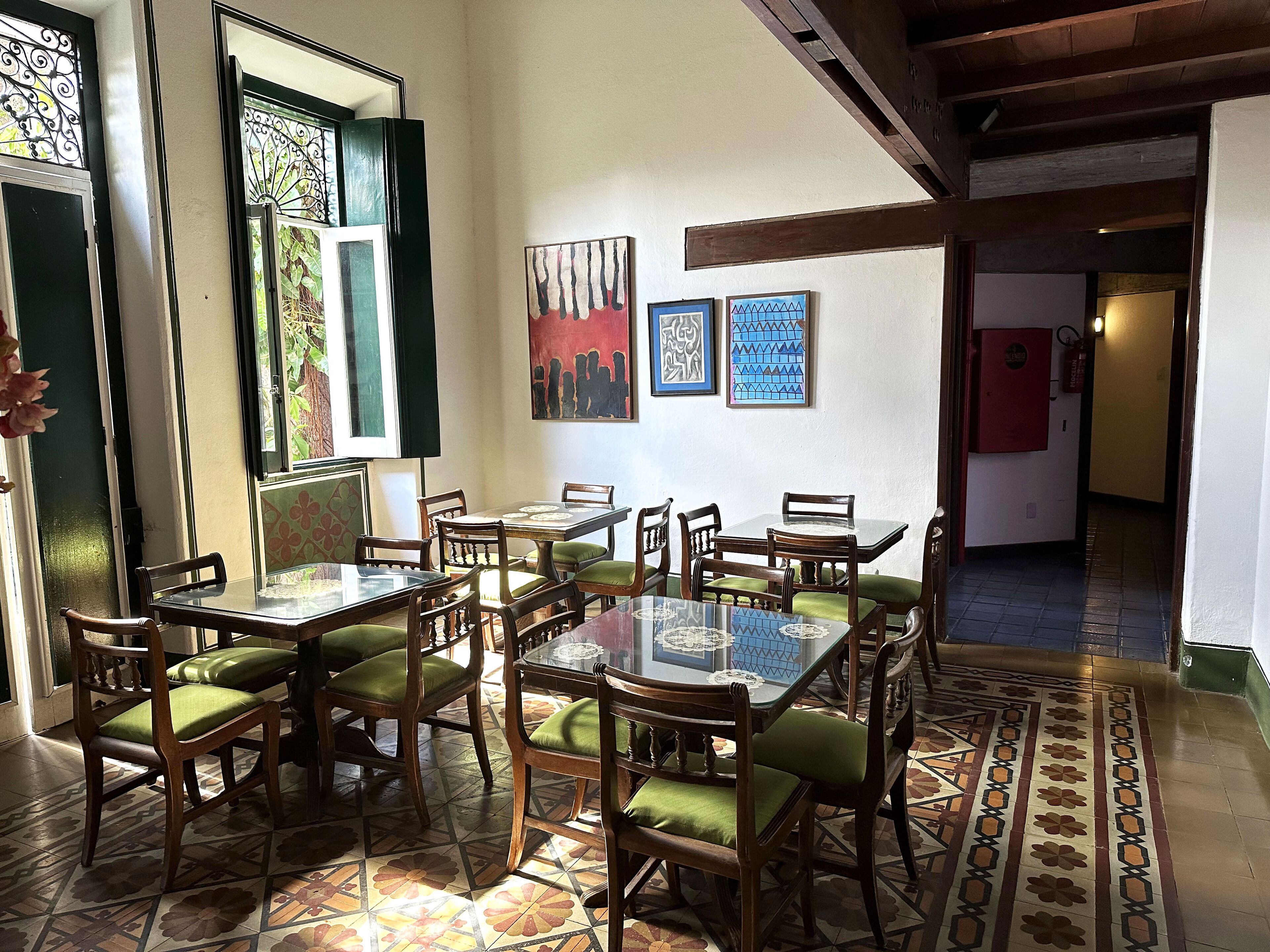 A dining area with a glass-top table surrounded by wooden chairs with green cushions, a tiled floor with a floral pattern, and artwork on the walls.