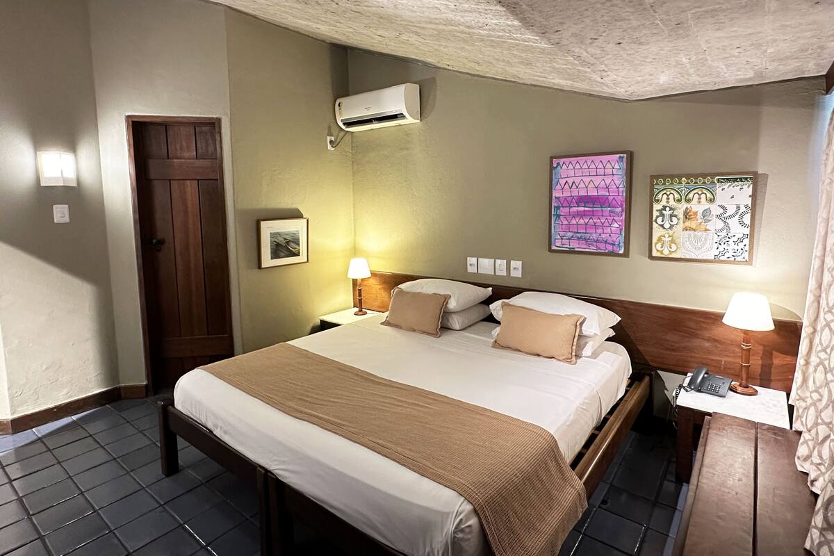 Habitación de hotel con cama, lámparas de noche, aire acondicionado, puerta de madera y un cuadro enmarcado en la pared.
