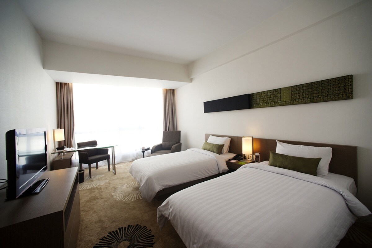 A hotel room with two beds, a desk with a TV, a chair, and a window with curtains.
